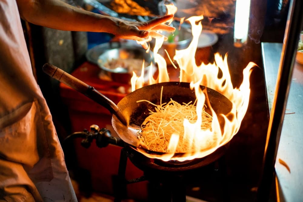 a street food chef tosses noodles into a wok while flames crawl up the side