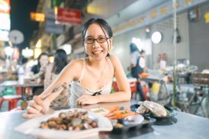 a young Asian woman with glasses eats seafood on a metal table on a Bangkok Street at night