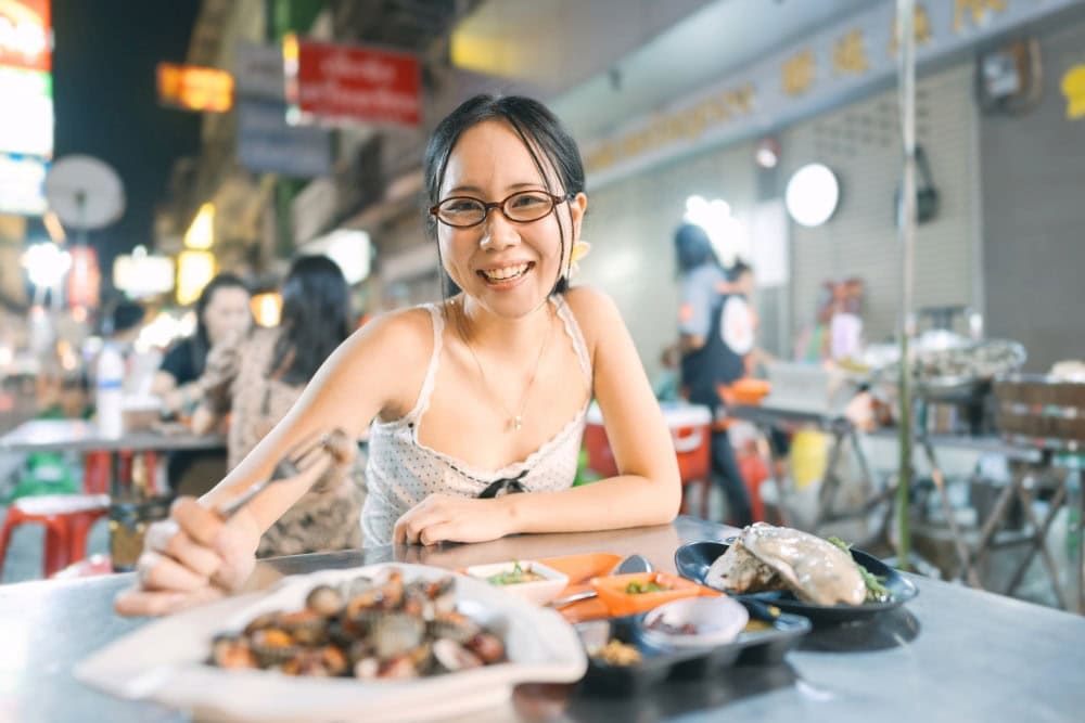 a young Asian woman with glasses eats seafood on a metal table on a Bangkok Street at night