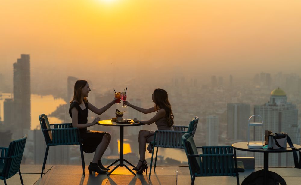 two women clink glasses while enjoying cocktails from a rooftop in Bangkok