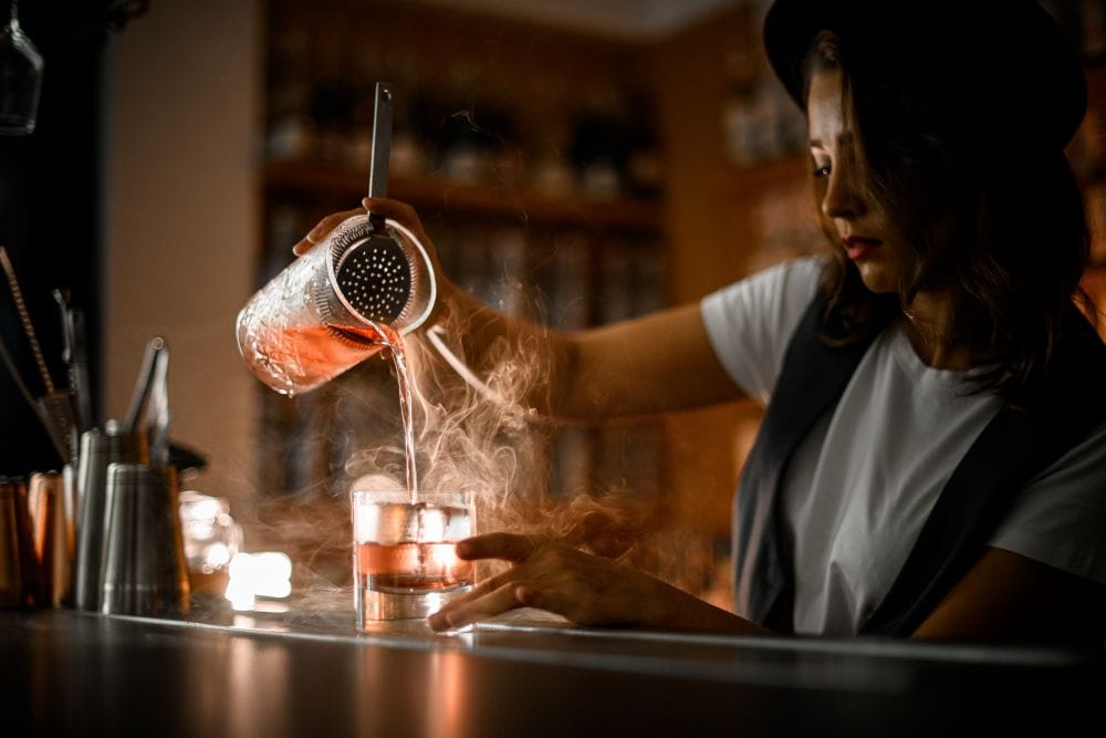 female bartender in a black vest pours a smokey cocktail at a speakeasy bar